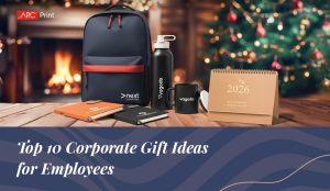 10 Corporate Gift Ideas for Employees including branded backpack, notebooks, water bottle, coffee mug, and 2026 desk calendar displayed on a wooden table with festive Christmas decorations and ARC Print logo — showcasing top corporate gift ideas for employees.