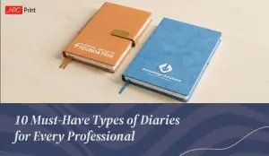 Two professional diaries on a light wooden surface – one in tan with 'Supreme Group of Foundation' logo and one in blue with 'ArrowEdge Solutions' logo. Text below reads '10 Must-Have Types of Diaries for Every Professional.